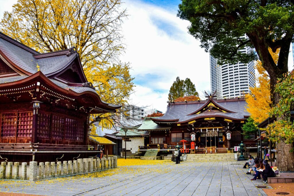 A vibrant photo capturing a traditional Japanese shrine with intricate wooden architecture, surrounded by autumnal yellow ginkgo trees, against a backdrop of modern skyscrapers under a clear blue sky.