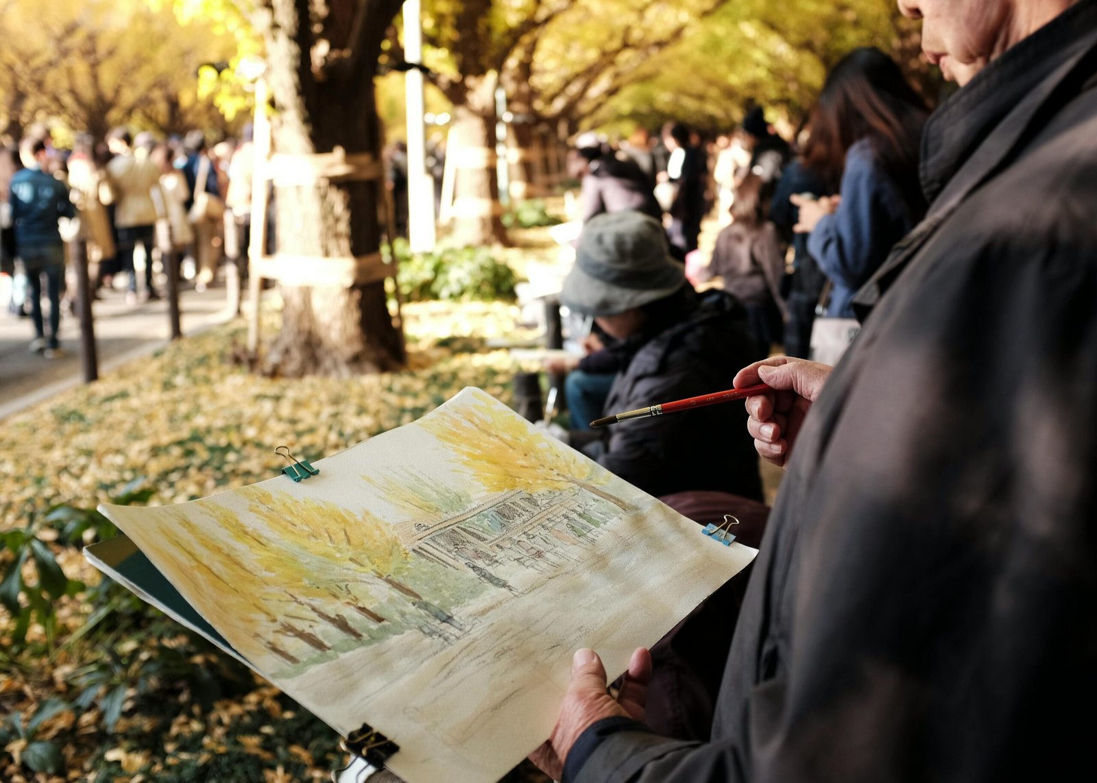 An artist painting a scene on a canvas in an outdoor setting with people walking in the background.