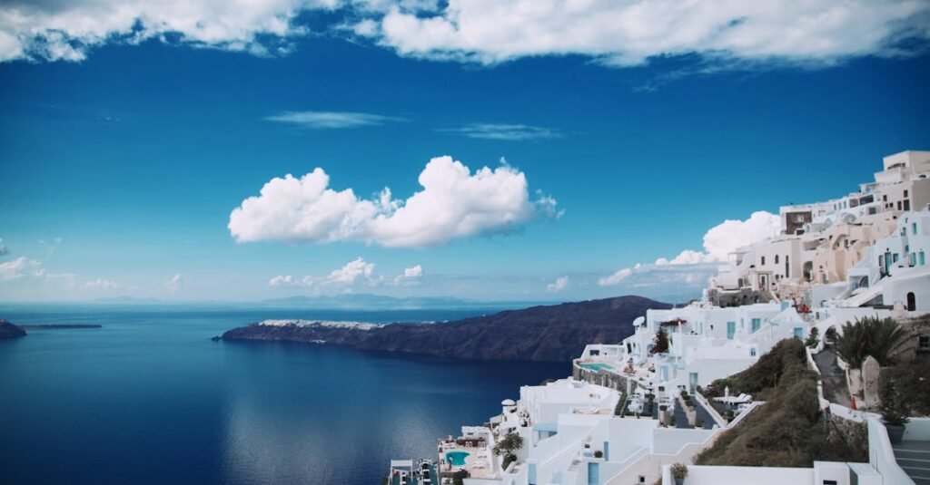 A panoramic view of Santorini, showcasing the iconic white buildings with blue accents perched on cliffs overlooking the Aegean Sea under a clear blue sky with scattered clouds.