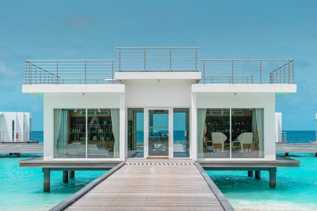 A modern overwater villa with a flat roof and large glass windows, featuring an oceanfront view under a clear blue sky.