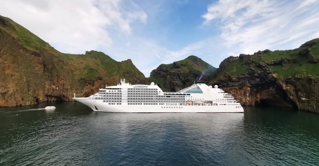 Luxury Seabourn cruise ship on a Caribbean and West Indies Vacation anchored in a serene bay with emerald waters and rugged cliffs under a clear blue sky.