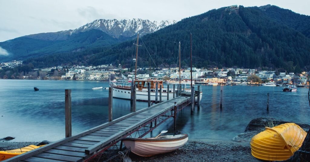 A serene view of a waterfront in Queenstown with a boat docked on a wooden pier, mountains in the background, and overturned yellow boats on the shore.