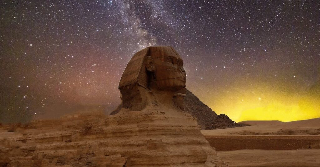 Great Sphinx of Giza illuminated at night with a starry sky and Great Pyramid in the background.