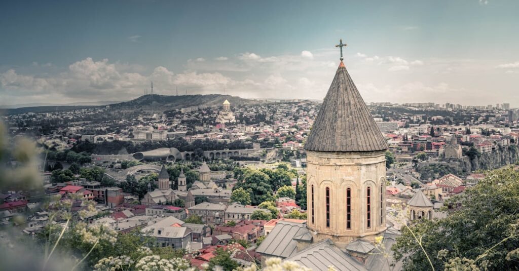 Panoramic view of Tbilisi with a prominent church in the foreground, modern buildings, and green areas spread across rolling hills.