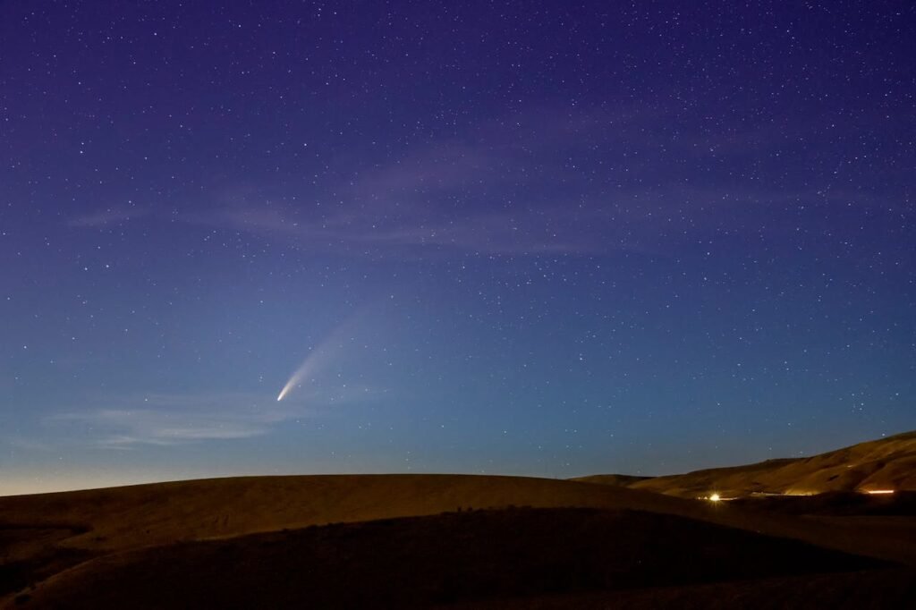 A comet streaking across a star-filled night sky above silhouetted rolling hills with distant lights on the horizon.