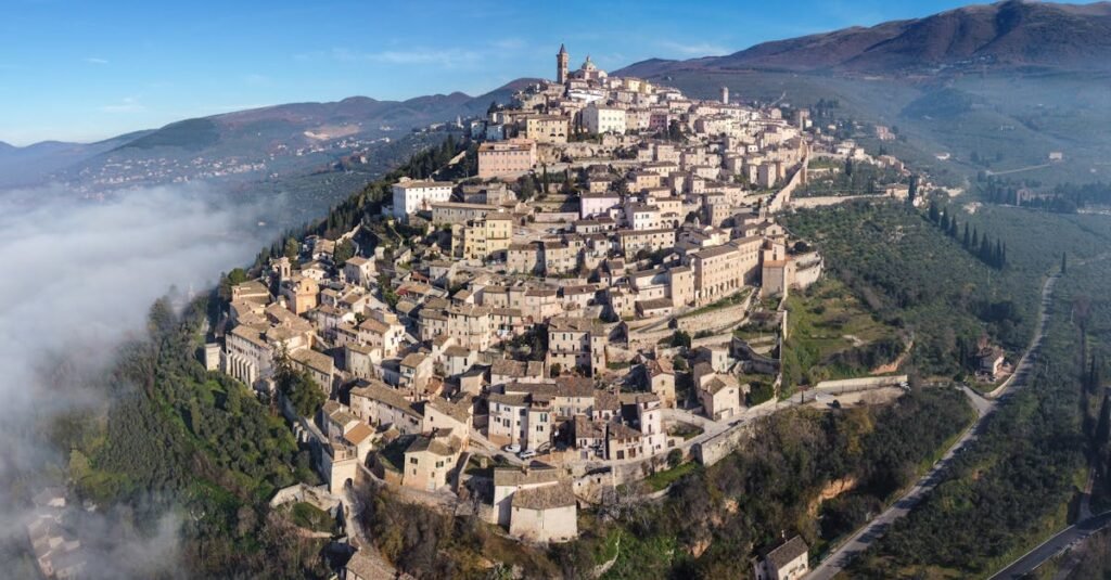 Aerial view of San Marino with historic buildings and fortifications perched on a hilltop, surrounded by lush greenery and low-lying clouds.