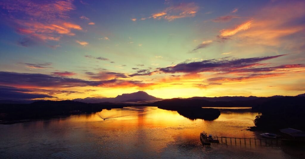 A vibrant sunset over a calm body of water with mountains in the background and a pier extending into the water.