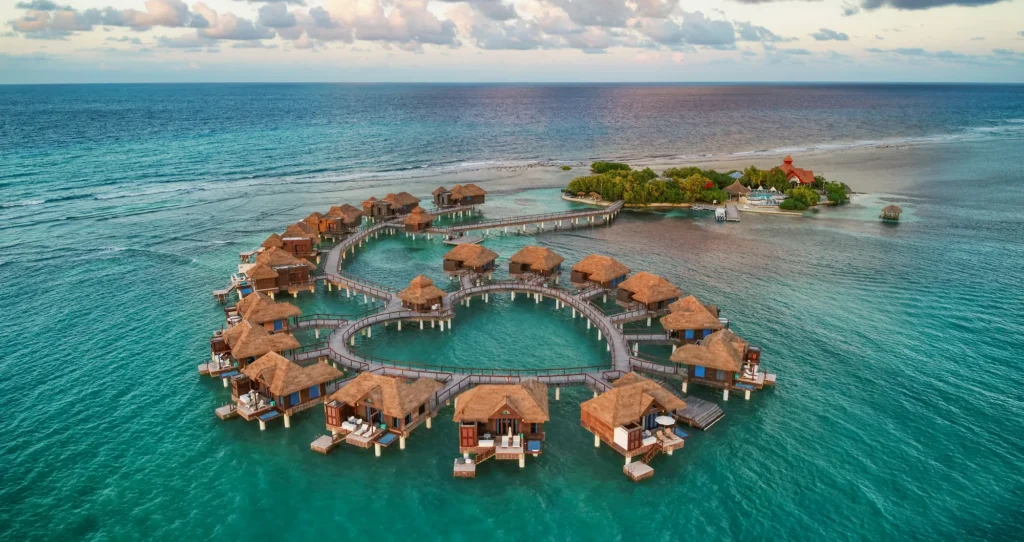 Aerial view of a heart-shaped arrangement of overwater bungalows with thatched roofs in a tropical setting, surrounded by clear turquoise waters.