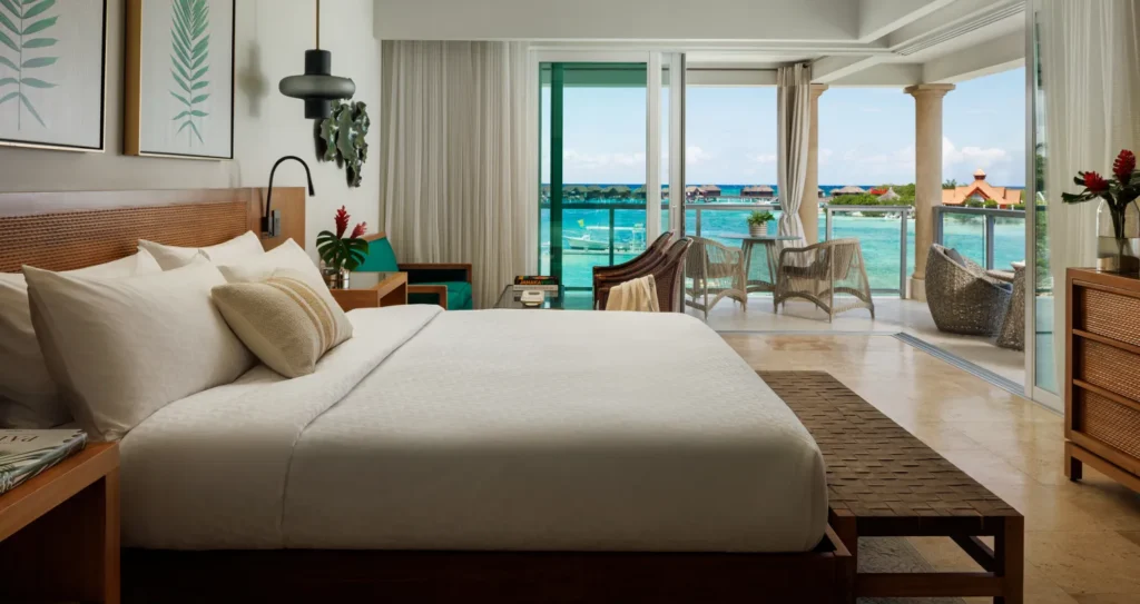 A luxurious hotel room with a large bed in the foreground, leading to an open balcony overlooking the sea.