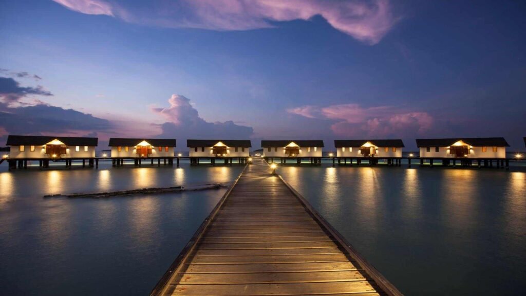 A serene twilight view of overwater bungalows extending from a wooden pier, with calm sea waters and a dramatic sky at dusk.