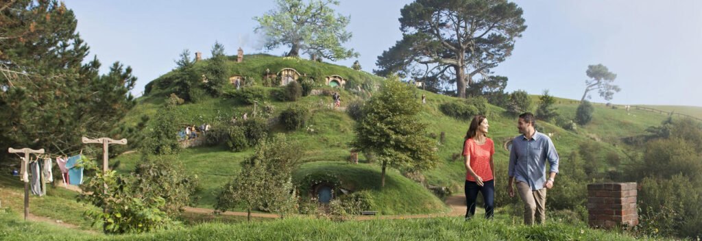 A scenic view of Hobbiton in Matamata, New Zealand, featuring lush green hills with hobbit holes and two people walking in the foreground.