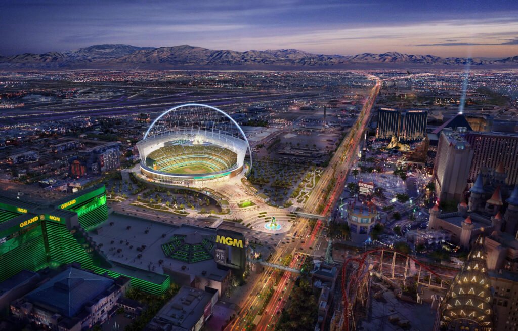 Aerial view of a modern domed stadium in an urban setting at dusk with surrounding cityscape and mountains in the background.