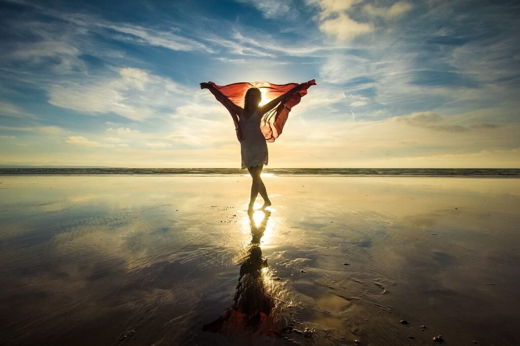 Person standing on a reflective beach at sunset, holding a flowing scarf, creating a silhouette.